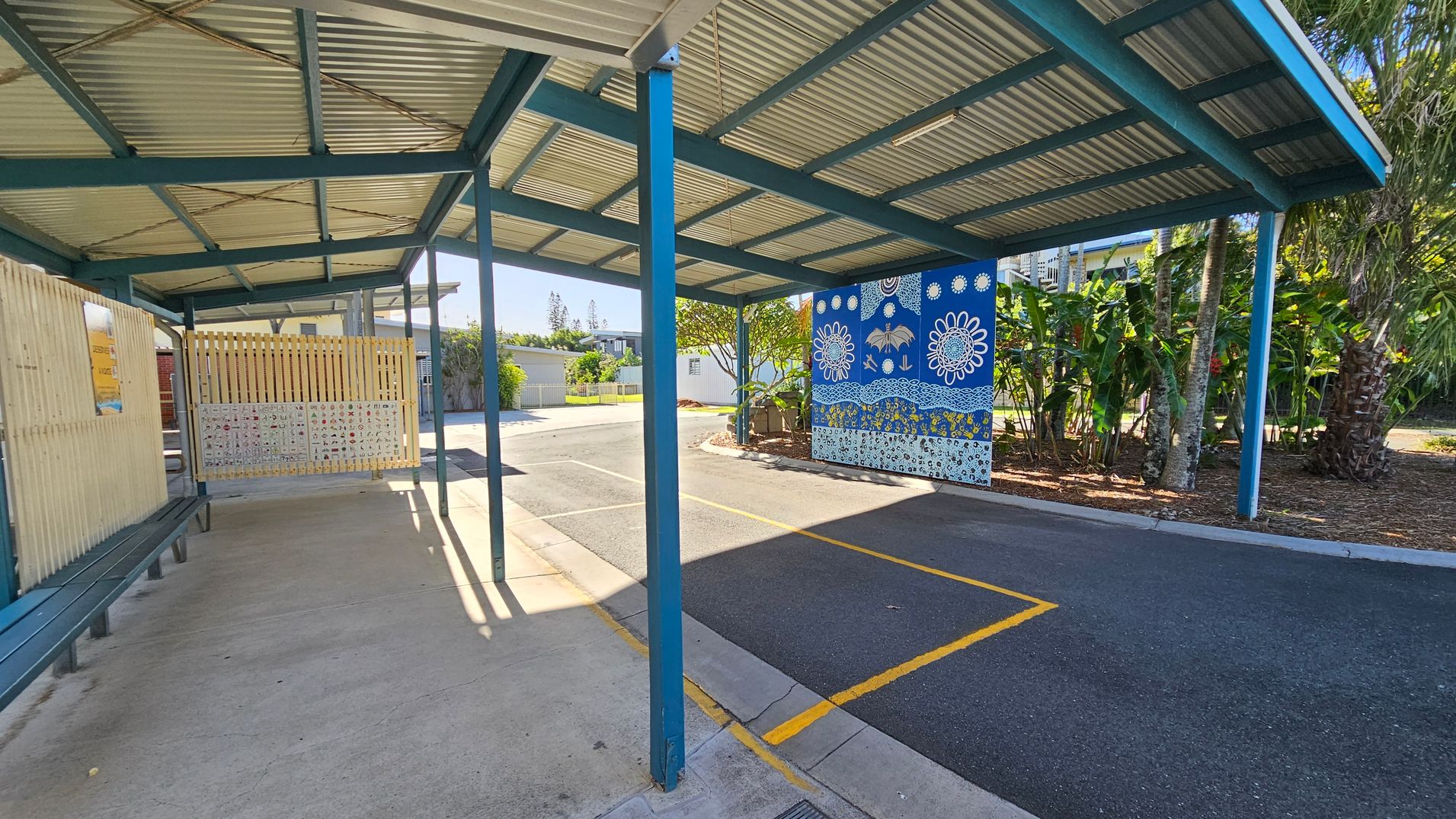 A covered walkway with a corrugated metal roof supported by blue beams. On the left, there is a wooden fence with a bench beneath it. The ground is a mix of concrete and asphalt. On the right, a vibrant blue mural with white floral and abstract designs is visible. Behind the mural, there are lush green plants and trees. The area is well-lit with sunlight casting shadows on the ground.