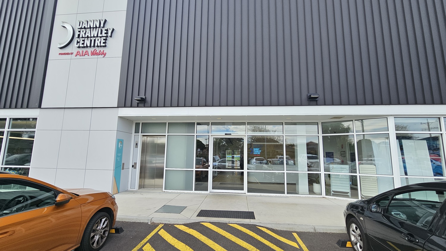 On the left, an orange car is partially visible. On the right, a black car is parked. In the middle, the entrance to the "Danny Frawley Centre" features large glass doors and windows, with a sign above the door. The background includes reflections of trees and other cars in the glass. The floor is a paved area with yellow-striped markings near the entrance.