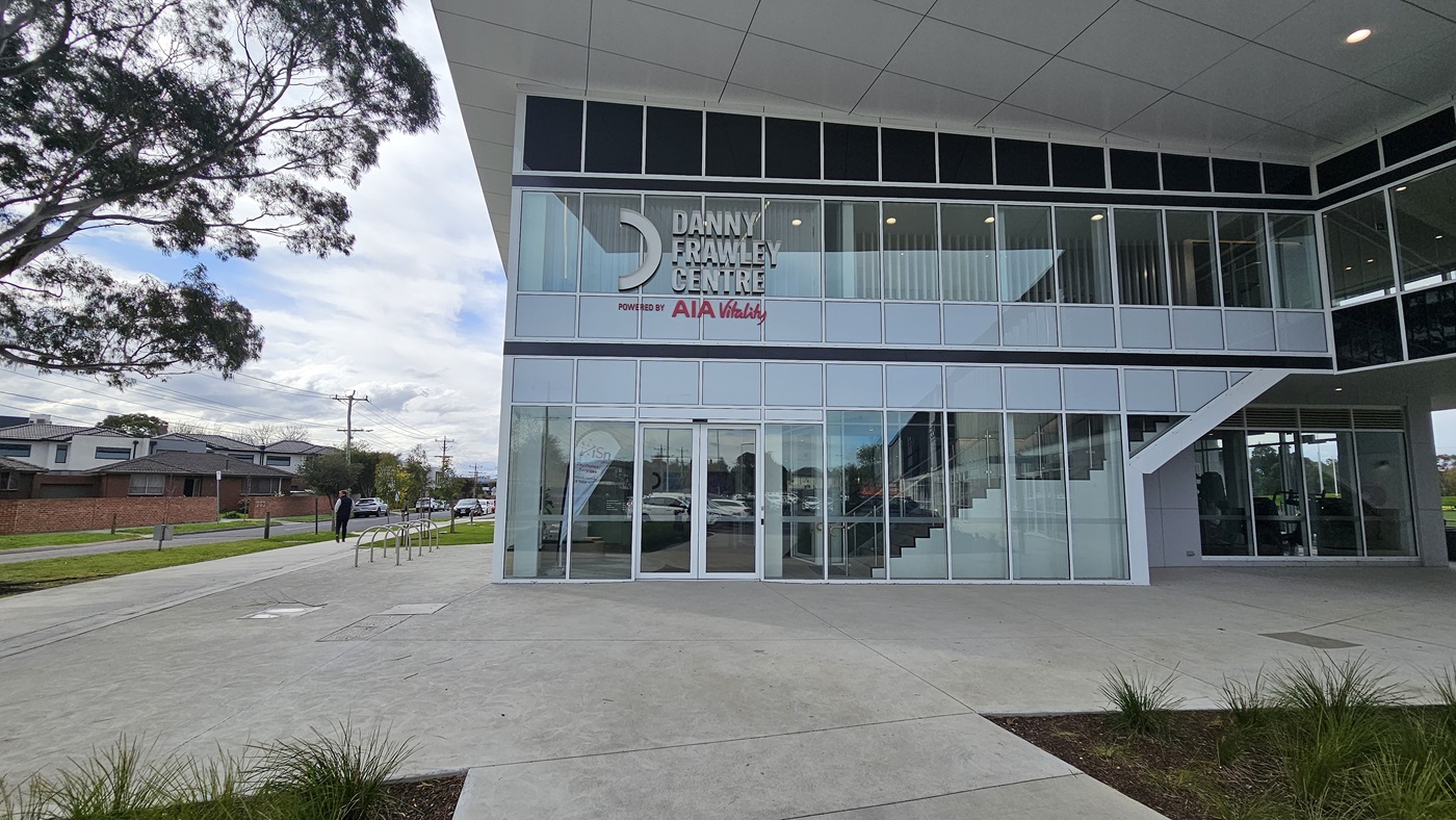 On the left, a large tree with spreading branches. On the right, a glass-walled section of the building with visible interior stairs. In the middle, the entrance to the "Danny Frawley Centre" with large glass doors and windows, and a sign above. The background includes a street with houses and a person walking. The foreground has a concrete pavement with some grass patches. The floor is paved, and the ceiling is a grid of white panels.