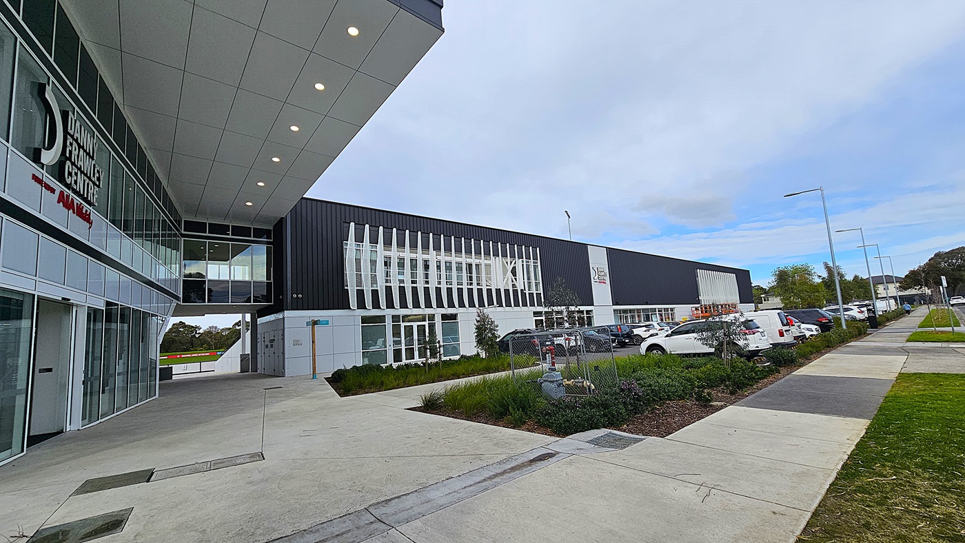 On the left, the "Danny Frawley Centre" sign is mounted on a glass-walled building with white framing. On the right, a long black building with vertical white louvres and several parked cars. In the middle, a covered walkway with a white ceiling featuring recessed lights. The background includes a row of street lamps and trees. The foreground has a concrete pavement with landscaped greenery and a small fenced area. The floor is paved, and the ceiling is a grid of white panels with lights.