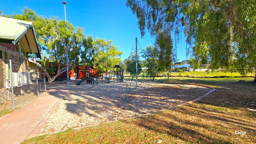 The child’s gym in Dayboro can be distinguished by a circular boundary on its edges and is right outside the chain-linked fence of Dayboro Pool and Gym. A part of the building shows a brown tiled wall. In the middle is an obstacle with green ropes, while to the back are playground climbers and slides. Further beyond is a building with a red wall, and to the right is a large grassland.