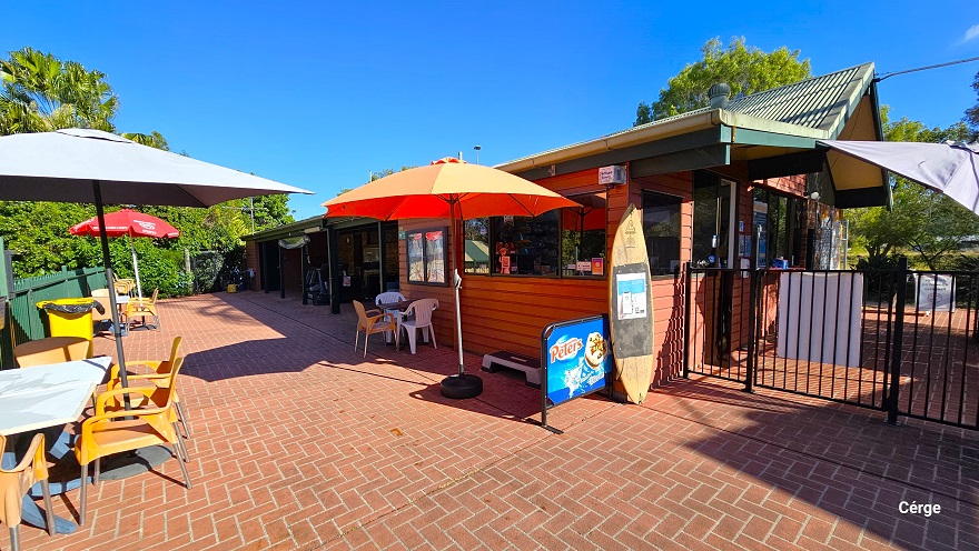 The outdoor seating area in Dayboro Pool & Gym has a natural vibe setting with an overlook of the tall trees with abundant foliage. There are glass windows on this side of the building, and it has a similar red brick floor to the reception. The white, orange, and red sun shades have orange, white plastic chairs, and a white table. A tall yellow garbage bin with a black garbage bag is placed against the green curvy-style fence. A wooden surfing board is left against the wall near the black fence.