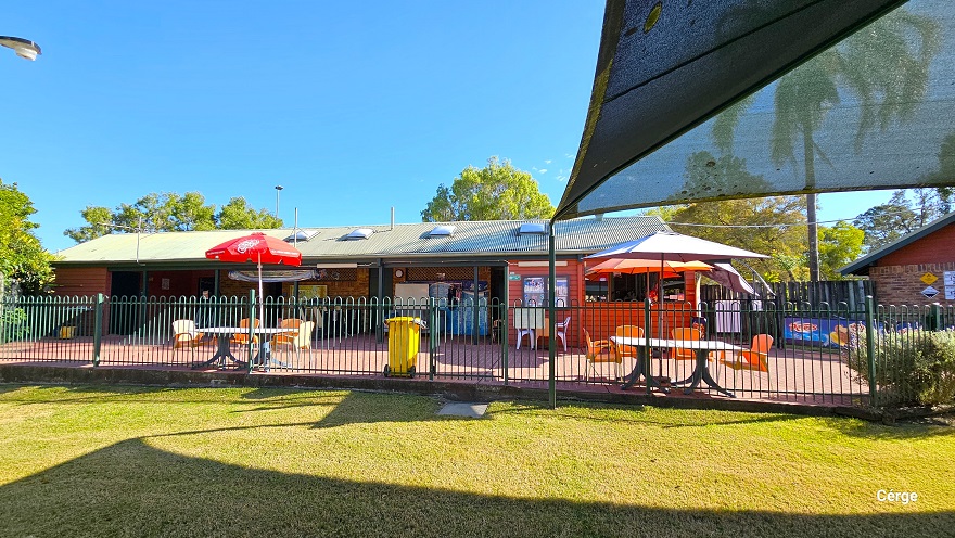 View of the seating area in front of the low-set 1970s-style red brick building when standing inside the green curvy-style fence. Within the green fence is an open area with a well-maintained height. This image also shows the corrugated roof of the building with square bumps.