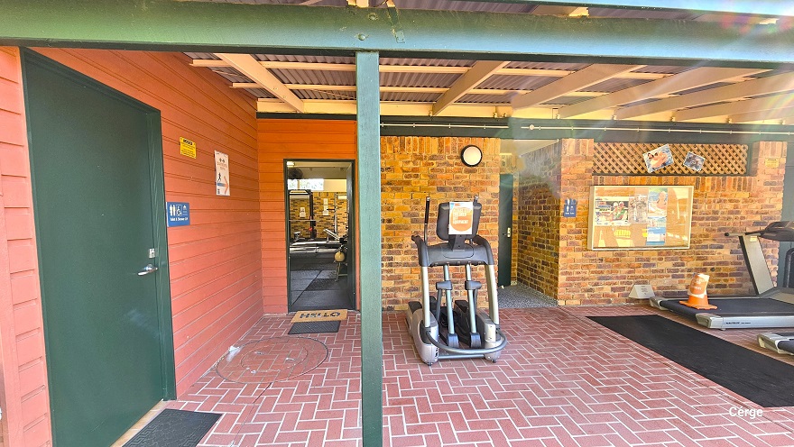 The 1970-style brick building with red corrugated walls and roofing has a red brick floor style. The building has green metal frames as support. Within the red corrugated wall is the accessible bathroom, which can be distinguished by the green door, a black mat, and a blue Unisex Toilet label. On the brick wall to the right is a painting and two photos. Two elliptical trainers are available in this area.