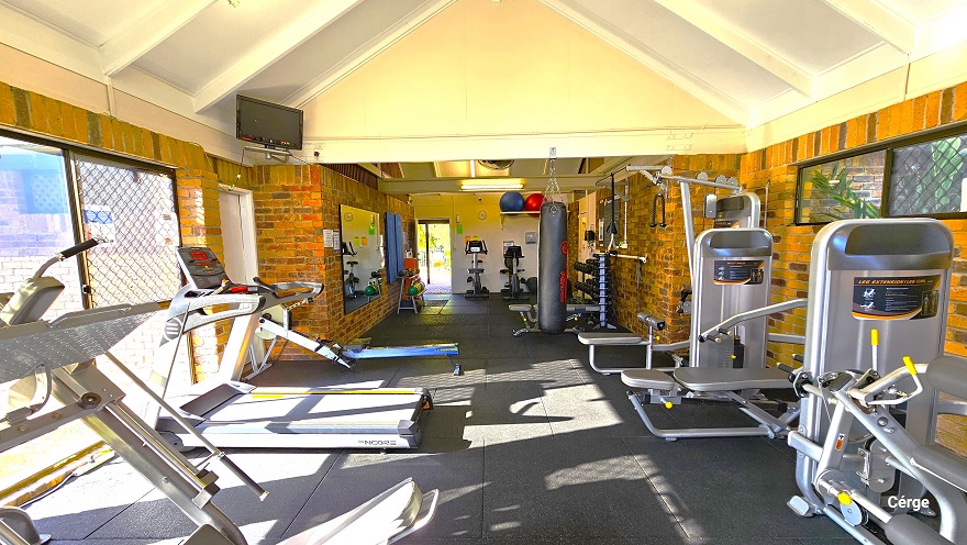 The image shows the back portion of the inside of Dayboro Gym. Mounted on the left wall is a small television above another white door. The glass windows reflect the silhouette of the chain-linked fence outside the building. On the upper left of the white wall next to the entrance door, there are blue and red yoga balls placed on a white rectangular frame. Two small glass windows can be found on the upper part of the right brick wall.