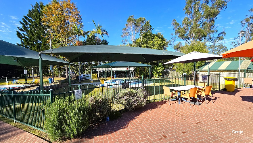 Red brick pathway leading to the six-lane, 25m lap outdoor pool. The pool is situated down a slight incline with a green sun shade on one end of the pool. A yellow sign board can be found under the green shade. To the left is a shed with a green corrugated wall and opening. Two green garbage bins with wheels are placed in front of the shed. To the right is another pool enclosed within a low green pool fence and is covered by two green sun shades.