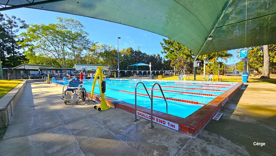 A 25m, 6-lane outdoor pool at Dayboro Pool and Gym with partial shading on one end. The pool has red-tiled edges all around with stainless steel pool ladders. A ‘Shallow end 1m’ and ‘No smoking’ sign is visible on the edge of the pool. There is an accessible aquatic wheelchair and a yellow pool hoist on the left pool deck. Each lane is separated by blue, red, and white floating rope lanes. Overhang the pool is strings of grey triangular flags wrapped around a pole on the opposite pool sides.