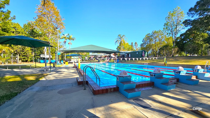 View from the diving platform of the 25m, 6-lane outdoor pool at Dayboro Pool and Gym. The pool deck has a rough surface floor to help avoid slipping. Each lane has a two-step stair with a grey non-slip cover blue diving platform. On top of the diving platform is a yellow semi-circle with a red caution on it. There is a green sunshade on the left with grey metal benches. Yellow floating devices are wrapped around poles with strings of blue triangular flags.
