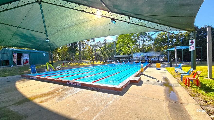 Different angle of the 25m, 6-lane outdoor pool at Dayboro Pool and Gym with partial shading on one end. Seen around the pool are yellow sign boards to remind guests of the do’s and dont’s of the pool. There is a small white sign for No Running and No Diving on a green post on the lawn to the right that has orange and blue monobloc plastic chairs and a grey bench.