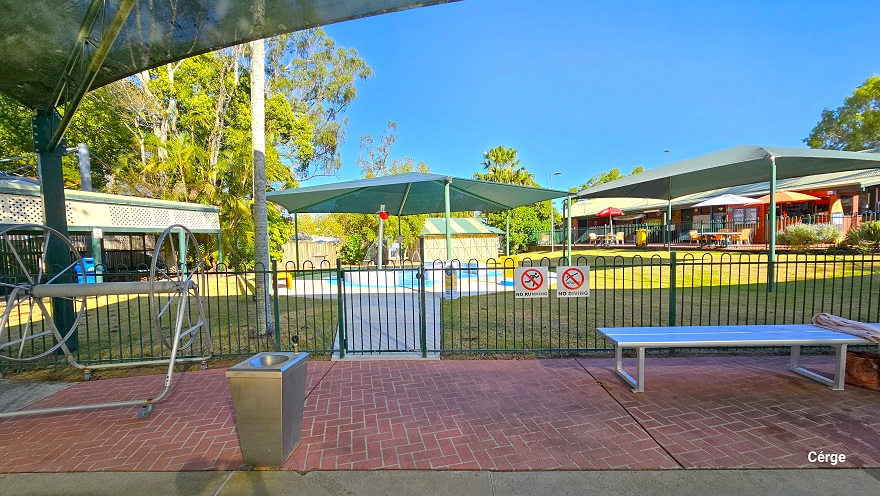 Red brick pathway next to the toddler splash pool enclosed by a green pool fence. Hanging on the green pool fence are the No Running and No Diving white signboards. There is a silver hose reel, a stainless steel wash basin, and a silver metal bench. Further to the left is a long sunshade with an upper side white cover that has several holes as designed.