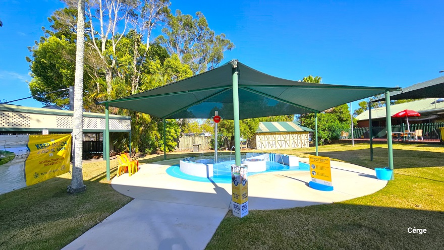 There is a smooth concrete pathway leading to the small toddler splash pool at Dayboro Pool & Gym. The pool is protected by a green sunshade wrapped around green posts. On a silver metal post is a large red lip graphic placed near the top of the post. The splash toddler aquatic activity offers interactive narrow water fountains from the two opposite white curves. There are yellow and red monoblock plastic chairs on the pool deck.