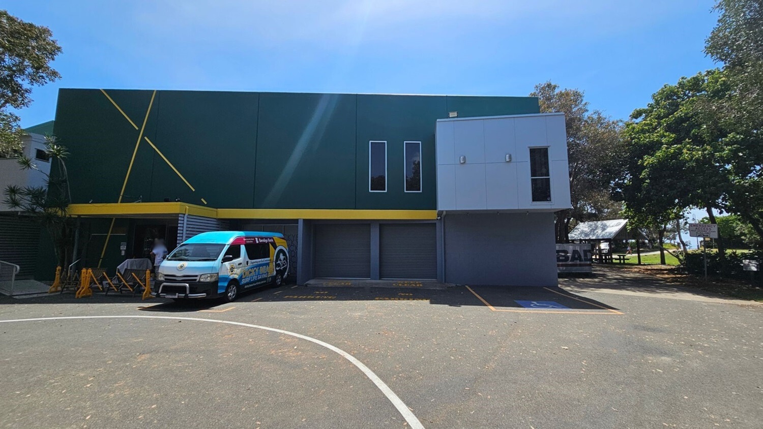 A two-storey building with a green and grey facade, featuring two narrow vertical windows on the upper level. A yellow stripe runs horizontally across the building. On the left, a van with colourful branding is parked in front of the building. Several yellow barriers are nearby. To the right, trees and a picnic area with a shelter are visible. The ground is covered with a textured surface, marked with a parking space.