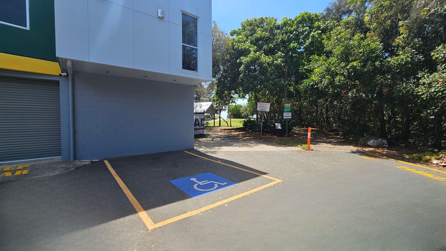 A parking area with a designated disabled parking space marked in blue and yellow on the ground. On the left, a building with a grey roller door and a yellow stripe above it. In the centre, a path leads to a grassy area with trees and a picnic table. Several signs are visible near the trees. The right side is bordered by dense greenery, and an orange bollard stands near the path. The ground is covered with scattered leaves.