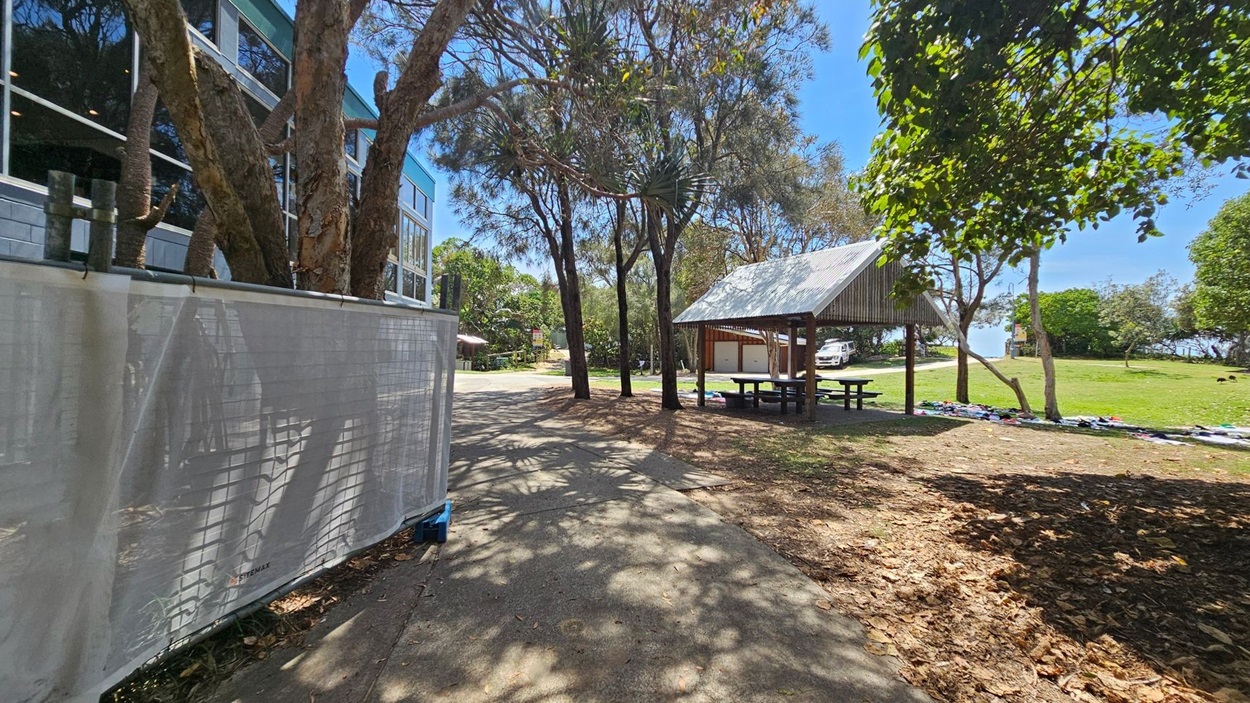 A paved path runs through a park area. On the left, there's a building with large windows and a temporary fence. Trees line the path, casting shadows on the ground. On the right, a small shelter with a metal roof covers picnic tables. Beyond the shelter, a grassy area extends with scattered leaves and a parked white vehicle. In the background, more trees and a glimpse of water are visible.