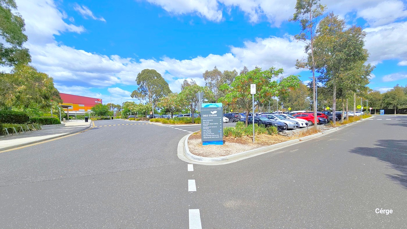 Car parking at Eagle Stadium. Upon arrival at the carpark, there is a grey signboard with a light blue water aesthetic background placed within the road verge for the different amenities with a corresponding arrow sign for its direction. On the right side of the grey signboard is a NO STOPPING OR NO STANDING SIGN, with the arrow pointing to the right. On the left side is a wide footpath with stainless steel bike racks.