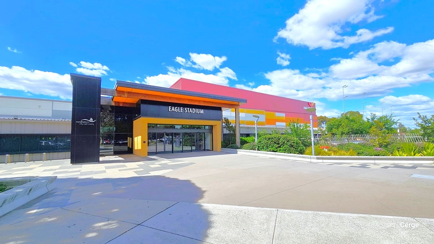 Eagle Stadium concourse with different floor styles. On either side of the concourse are alternating dark and light grey tiles, while in the middle is a smooth cemented floor connected to the automatic, glass sliding doors with black metal frames. On the left side of the floor-to-ceiling glass window is a black tile building column with the Wyndham City logo and slogan: city.coast.country. The name of the facility is detailed on the black wall above the entrance doors.