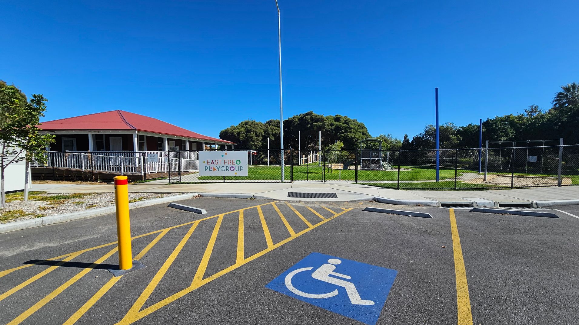 A parking area with a marked disabled parking space in the foreground, featuring a blue wheelchair symbol. Yellow lines and a yellow bollard are visible. In the background, a building with a red roof and a white railing is on the left. A sign reading "East Freo Playgroup" is attached to a fence, with a grassy area and playground equipment behind it. Trees are visible in the distance.