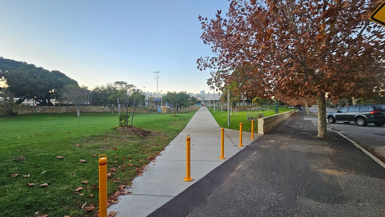 A pathway extends into the distance, flanked by green grass on the left and a row of trees with brown leaves on the right. Yellow bollards line the path's entrance. To the right, a parked car is visible on the road. In the background, there are more trees and a building with a playground. The sky is clear, and fallen leaves are scattered on the ground.