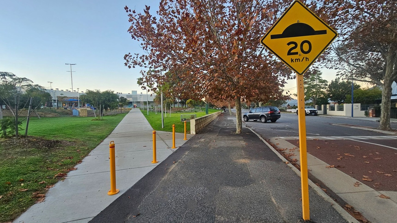 A pathway runs through the centre, bordered by a grassy area on the left and a road on the right. A yellow speed bump sign indicating "20 km/h" is prominently displayed on the right. Trees with brown leaves line the path, and a few cars are parked along the road. Yellow bollards are placed along the path, and a building is visible in the background. Fallen leaves are scattered on the ground.