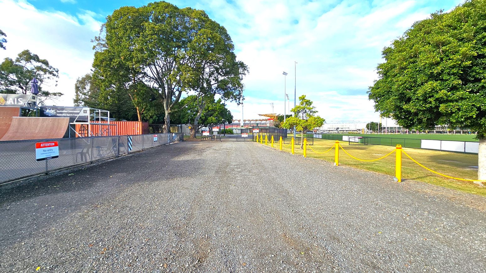 A gravel path runs through the centre, flanked by trees on both sides. On the left, there's a fenced area with a skate ramp and a sign that reads "Attention No entry." On the right, a grassy field is bordered by a yellow rope barrier. In the background, there are more trees and a sports field with floodlights. The sky is partly cloudy.