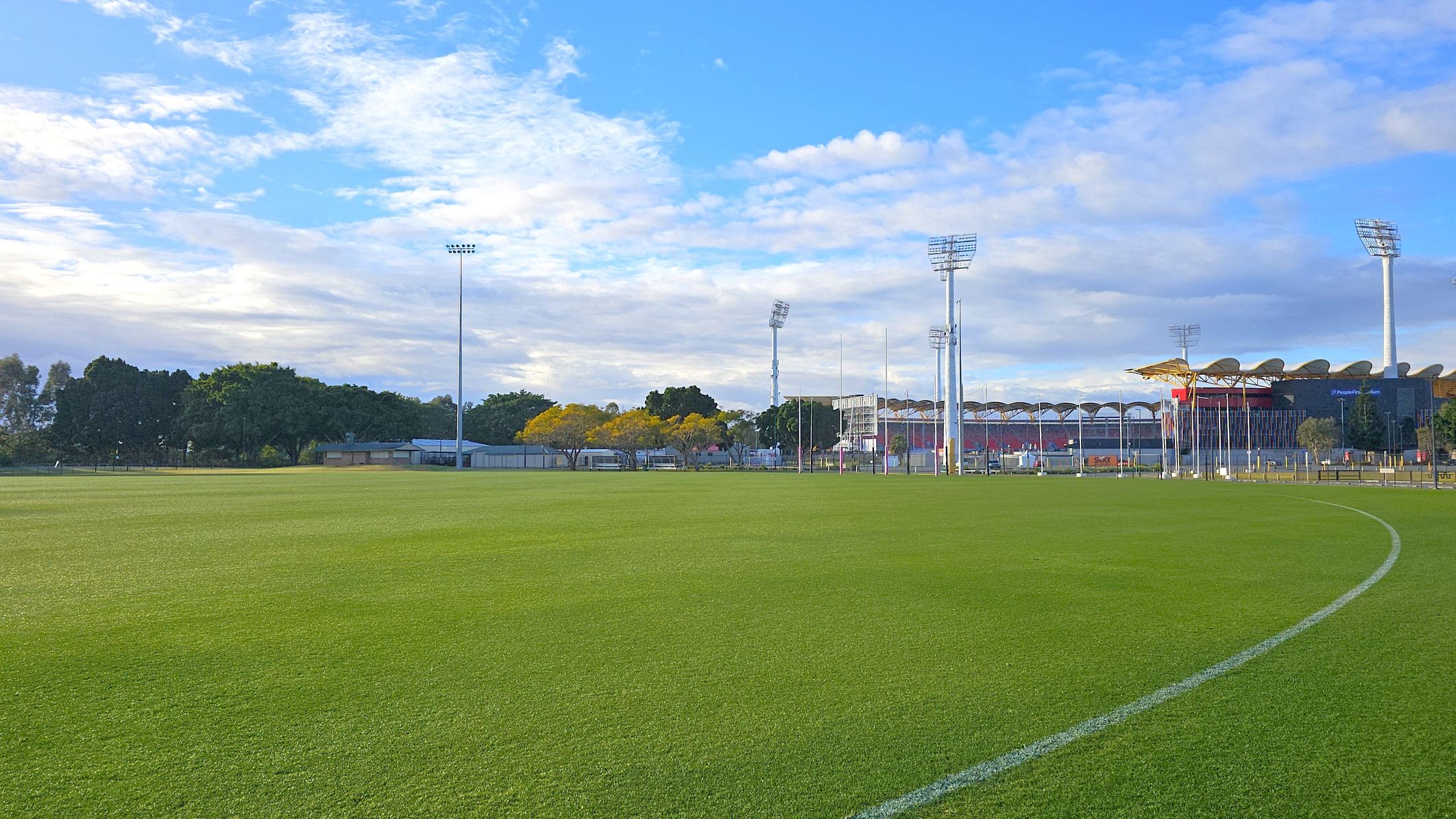 A large, well-maintained grassy sports field with a white boundary line curving across the foreground. In the background, there are tall floodlights and a stadium with a distinctive roof design. Trees with green and yellow foliage are visible to the left, along with some small buildings. The sky is partly cloudy with patches of blue.