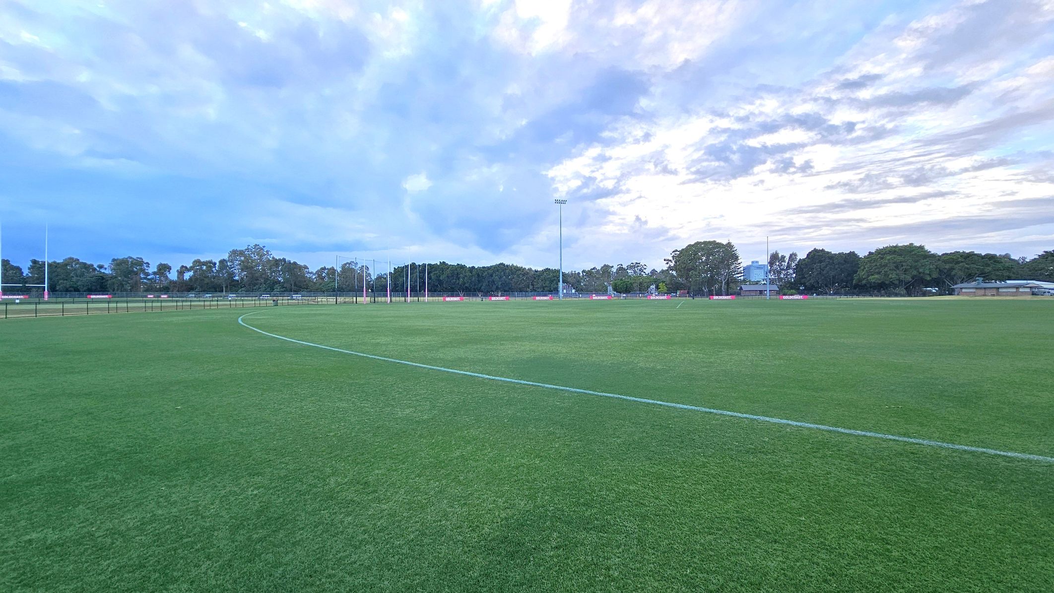 A wide, open sports field with well-maintained green grass. In the background, there are several tall light poles and a row of trees lining the perimeter. To the right, a few buildings are partially visible among the trees. The sky is overcast with a mix of blue and grey clouds.
