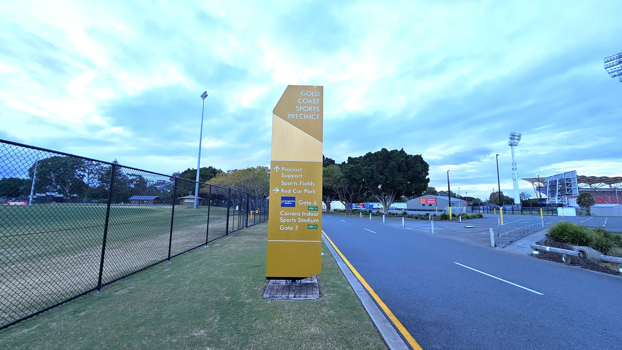 A tall, yellow sign stands on a grassy area, displaying directions for the Gold Coast Sports Precinct. To the left, a chain-link fence encloses a sports field with trees in the background. On the right, a paved road runs alongside the sign, leading towards a stadium with visible floodlights and seating. The area is bordered by trees and some low buildings.