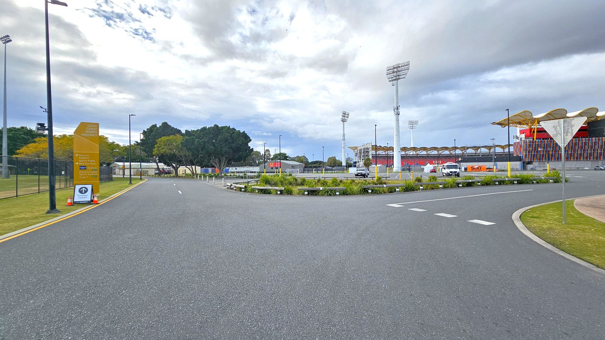 A wide road curves to the right, bordered by a grassy area with a few small plants. On the left, a tall yellow sign reads "Gold Coast Sports Precinct" with additional information below. Several tall floodlights are visible in the background, along with a large stadium structure featuring colourful vertical panels. A few vehicles are parked near the stadium, and trees are scattered throughout the area. The sky is overcast.