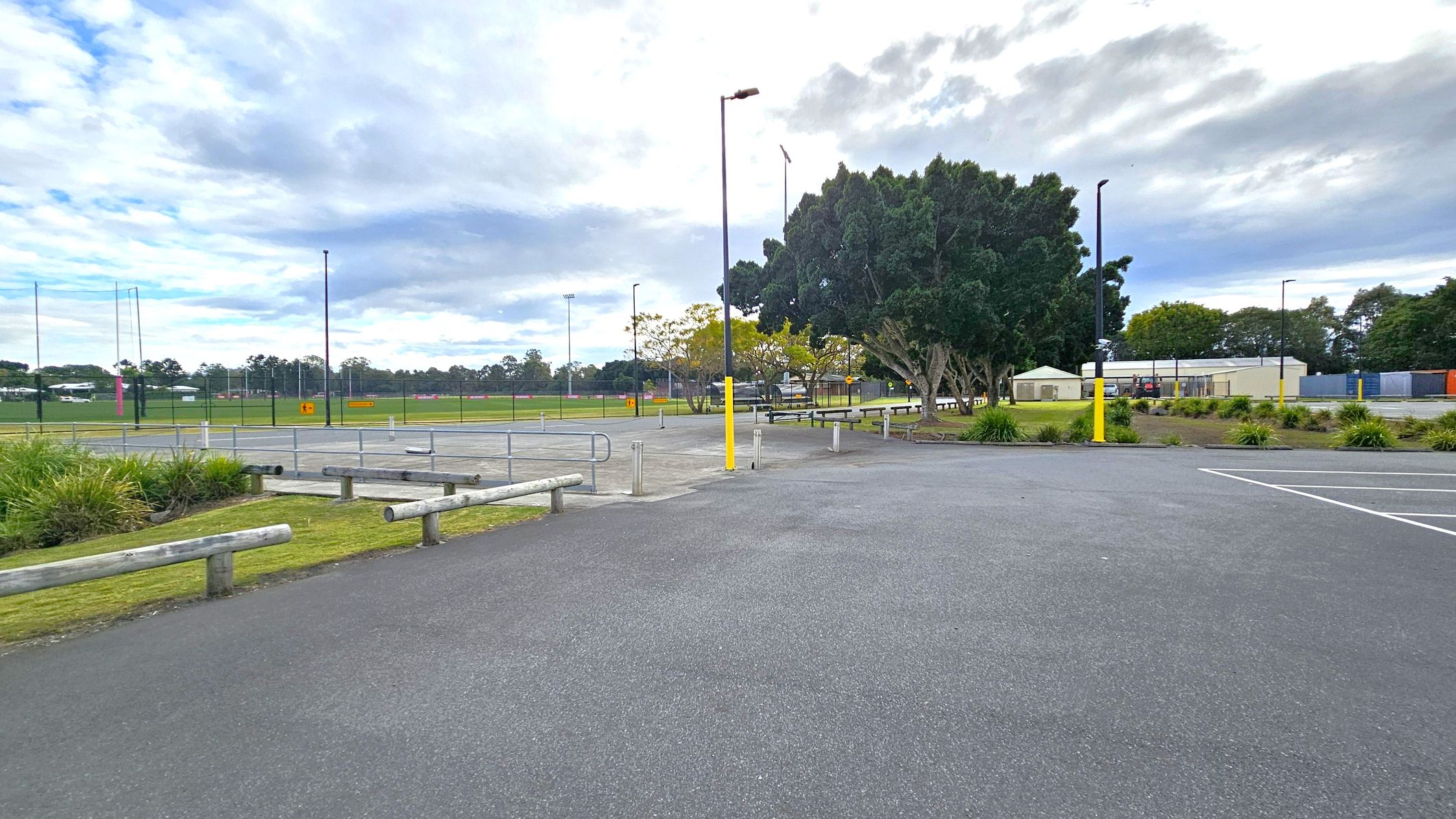 A paved area with parking spaces in the foreground, marked by white lines. Wooden barriers and small patches of grass with shrubs are on the left. In the middle, there are several tall lamp posts with yellow bases. The background features a sports field enclosed by a fence, with goalposts visible. To the right, there are trees and a few small buildings, possibly storage units or sheds. The sky is overcast with clouds.