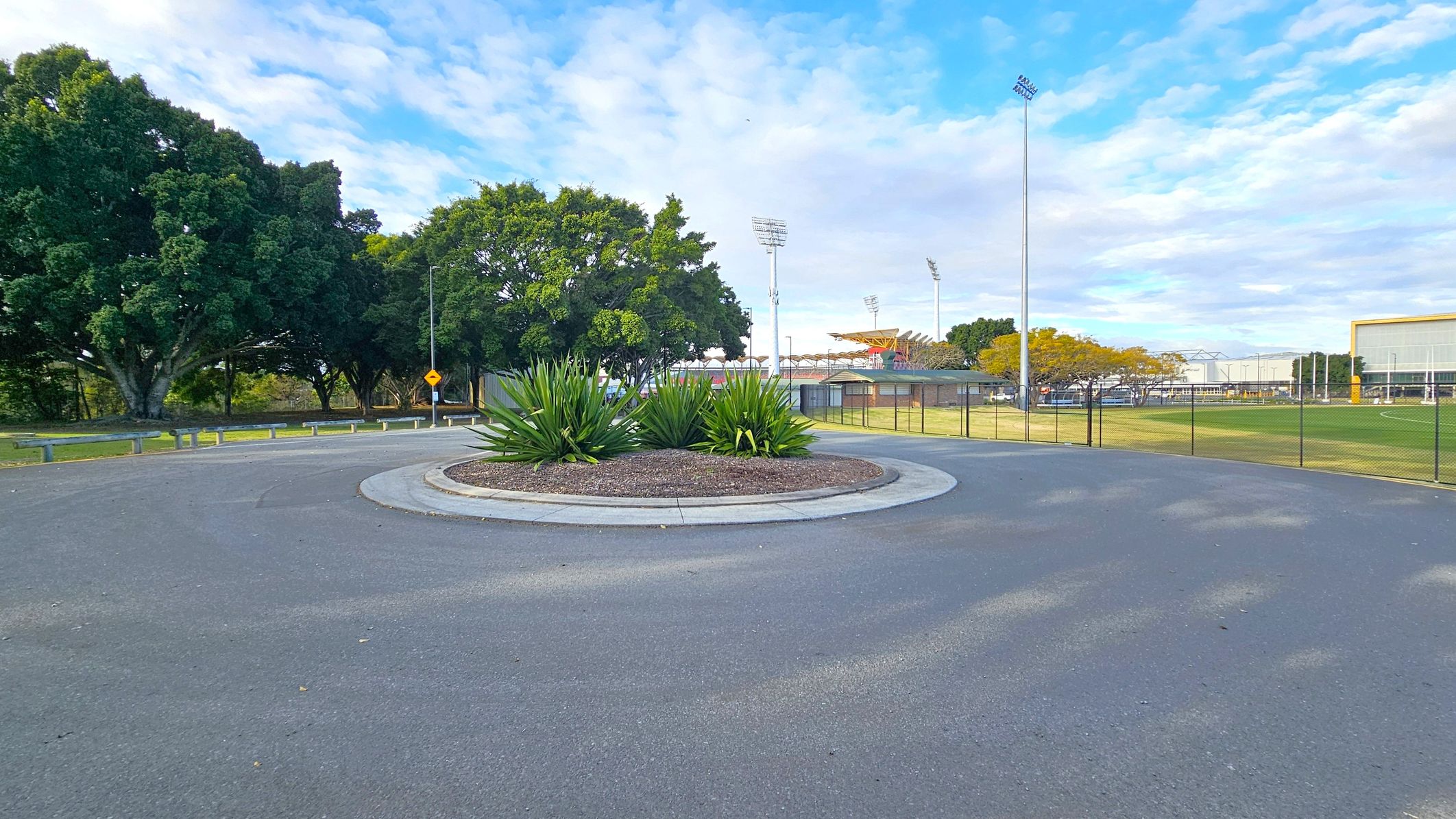 A roundabout with a central garden of green plants and mulch is in the foreground. To the left, there are large, leafy trees and a road with metal barriers. In the background, tall stadium lights and a sports field with a fence are visible. On the right, a building with a yellow trim and a grassy area can be seen. The sky is partly cloudy.