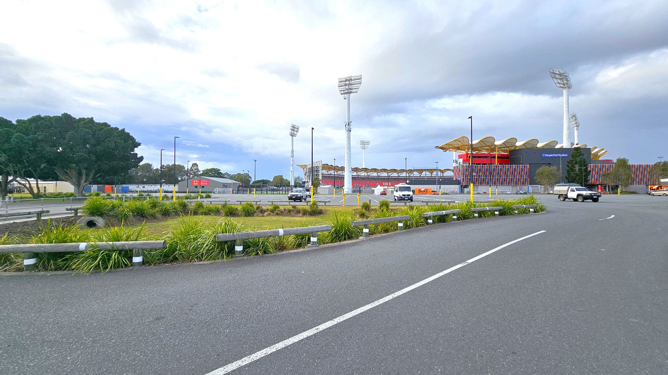 A large stadium with colourful vertical panels and a wavy roof is on the right, with tall floodlights surrounding it. In the foreground, there is a paved road with white lines and a grassy area with low bushes and a metal guardrail. Several vehicles, including a white van and a black car, are parked near the stadium. On the left, there are large trees and some industrial buildings with signage. The sky is overcast.