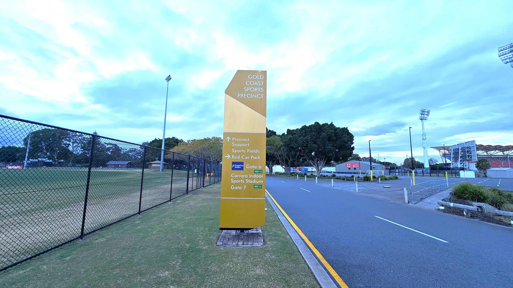 A tall, yellow sign stands in the centre, displaying directions for the Gold Coast Sports Precinct. On the left, a chain-link fence borders a grassy sports field with trees and a tall floodlight. On the right, a paved road with a yellow line runs alongside a car park and a stadium with a distinctive roof structure. Trees and a few buildings are visible in the background.