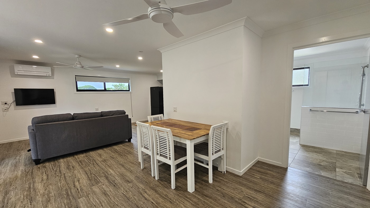 Inside the cabin, timber floors complement white walls and ceilings adorned with a ceiling fan and LED lights. A grey couch sits to the left, alongside a wooden dining set. A flatscreen TV and air conditioning are mounted on the left wall. Next to them, a glass window offers natural light, with a fridge nearby. To the right, a grey door leads to the bathroom, showcasing a shower with handgrabs, white walls, and grey tiles.