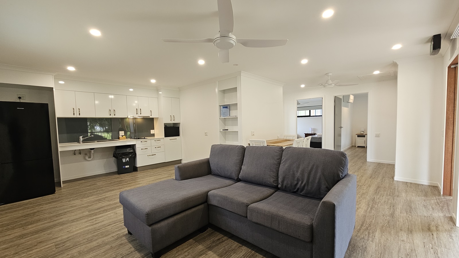 In the centre is an L-shaped grey couch. To the left is the kitchen area, equipped with cabinets, sink, drawers, microwave oven, and a fridge. Adjacent to the couch is a dining table. To the right, a door leads to the bedroom. The floor is timber, with white walls and ceiling featuring LED lights and ceiling fans.