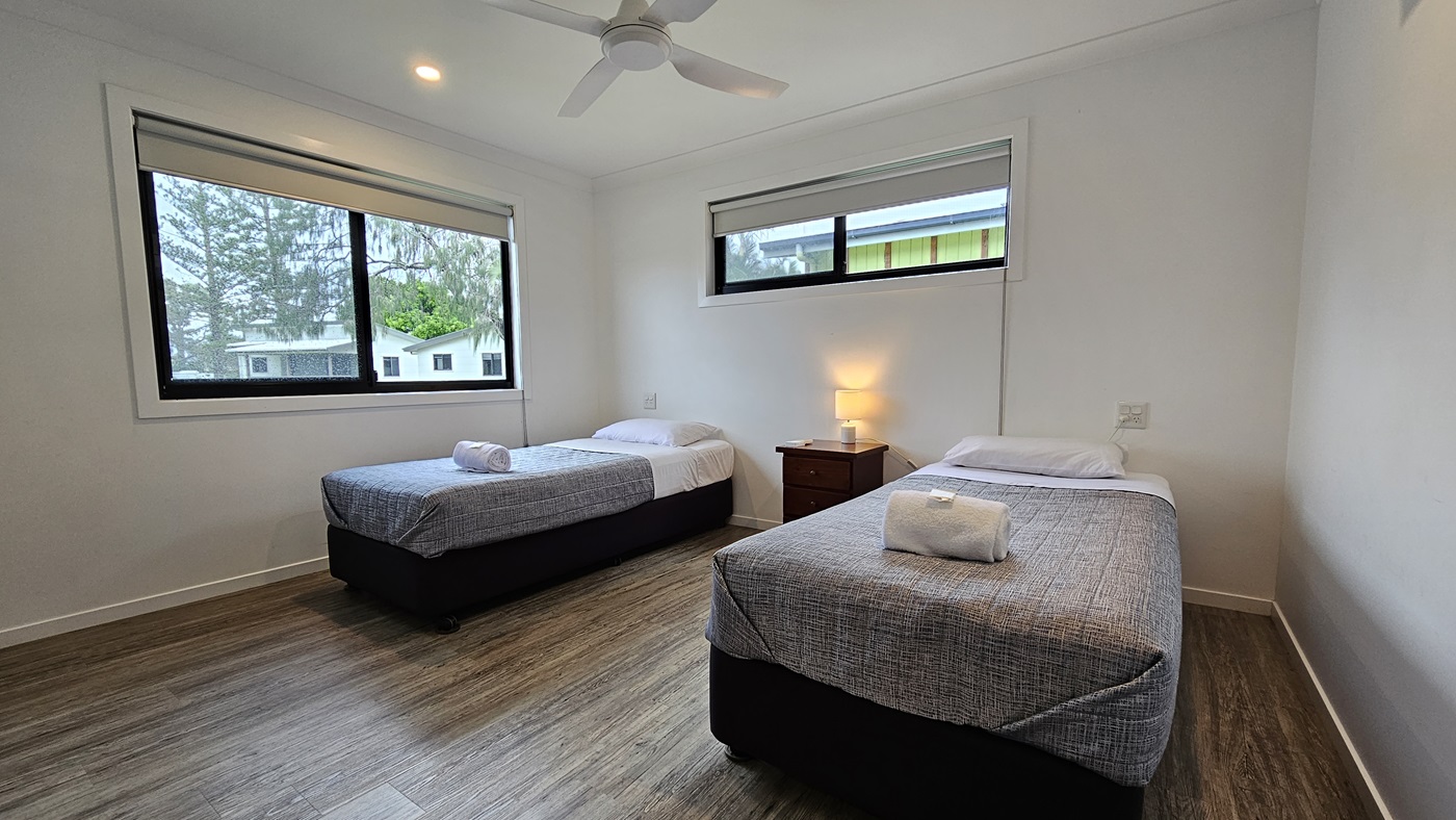 In the room, two single beds with grey covers stand opposite each other. White pillows and a neatly rolled white towel sit atop each bed. A window is positioned on the middle and right walls. Between the beds, a bedside table hosts a lampshade. The walls and ceiling are painted white, illuminated by LED lights and a ceiling fan. The floor is timber. Outside the window, trees and other cabins are visible.