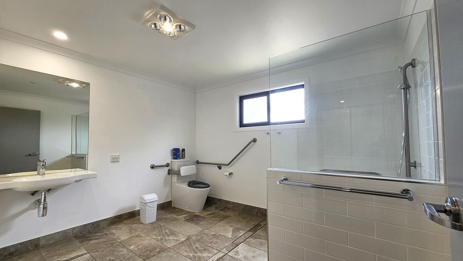 In the accessible bathroom, a toilet bowl with hand grabs is situated in the left corner, accompanied by a rubbish bin and a tissue dispenser. Adjacent to this is a hand basin with a mirror mounted on the glass wall. To the right, a shower is present, separated by a combination of glass and grey brick tiles. The floor features grey tiles with a marble design. The walls are white, and the ceiling is white with LED lights.