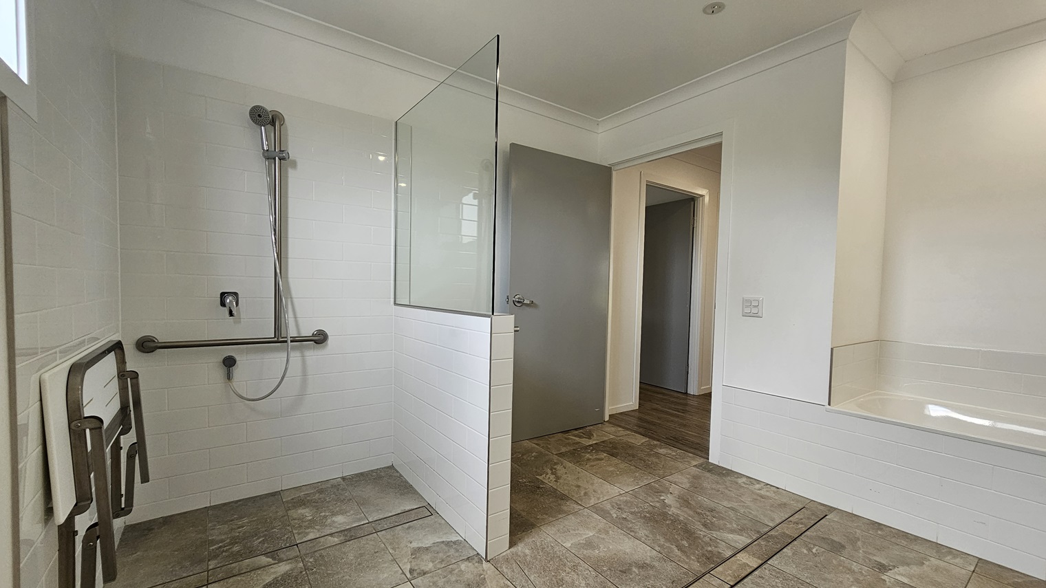 In the accessible bathroom, a shower with a handrail and foldable bench is on the left, separated by a partition of glass and grey brick tiles. On the right is a bath tub against the wall. The floor boasts grey tiles with a marble design. The walls and the ceiling are white with LED lights.