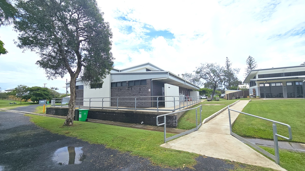 In a grassy area stands a low-rise facility with a grey brick facade. A brick pathway leads to it, alongside a step-free ramp with railings. Four gas tanks are displayed in front of the building. To the left is a round circular structure, and to the right is a wooden planter box with plants. Trees surround the area.