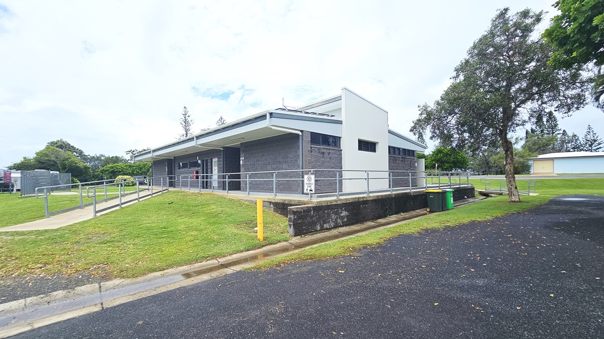 In the middle stands a low-rise facility with a dark grey brick facade. An accessible ramp entry with handrails is on the left, with an asphalt road to the right. Two rubbish bins are placed near the concrete wall. Surrounding the area are trees, grass, and other visible building structures in the distance.