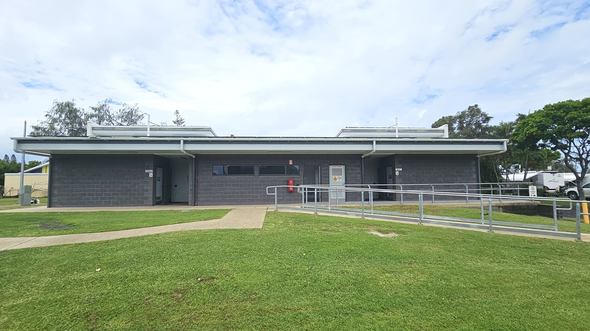 In the middle stands a low-rise facility with a dark grey brick facade, featuring small windows and multiple doors. An accessible ramp entry with handrails is on the right. To the left, brick pathways cut through the grass and lead to the building. Surrounding the area are trees, grass, and other visible building structures in the distance.