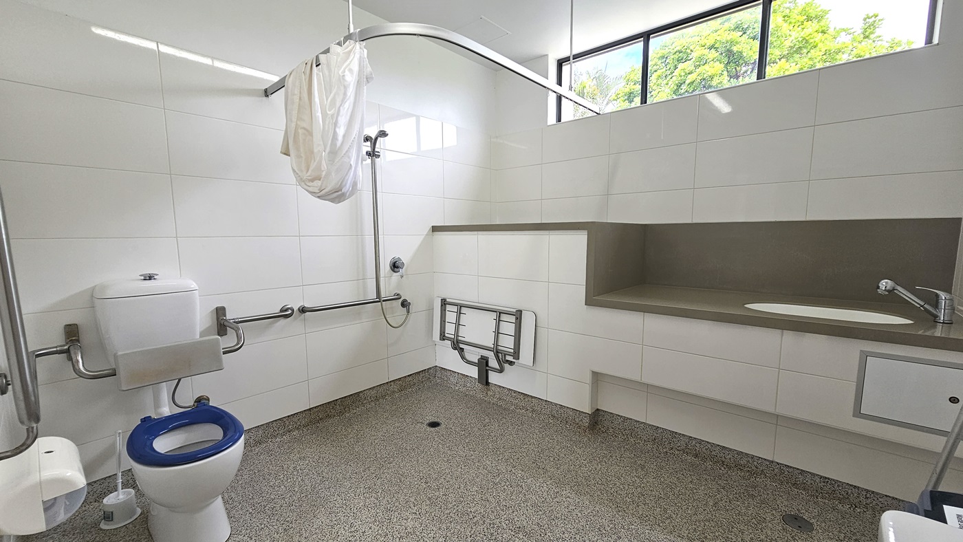 Inside the accessible bathroom, a toilet bowl with hand grabs, a tissue dispenser, and a toilet brush are on the left. On the right, a shower with handrails and curtain, alongside a foldable bench chair mounted to the wall. Adjacent, is a lavatory with a sink. The walls feature white tiles with glass windows. Floor tiles have a granite design.