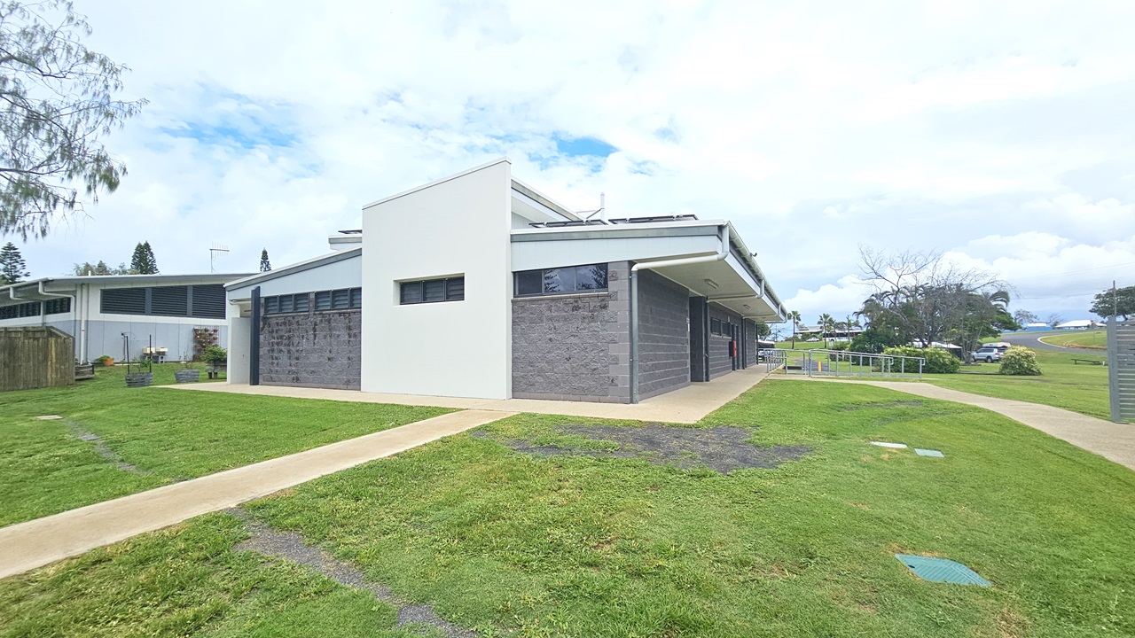 In the middle stands a low-rise facility with a dark grey brick facade, featuring small windows. An accessible ramp entry with handrails is on the right. Brick pathways cut through the grass, leading to the building. To the left is another building structure and planter boxes. Surrounding the area are trees and grass. Vehicles are parked under the shade of trees visible in the distance.