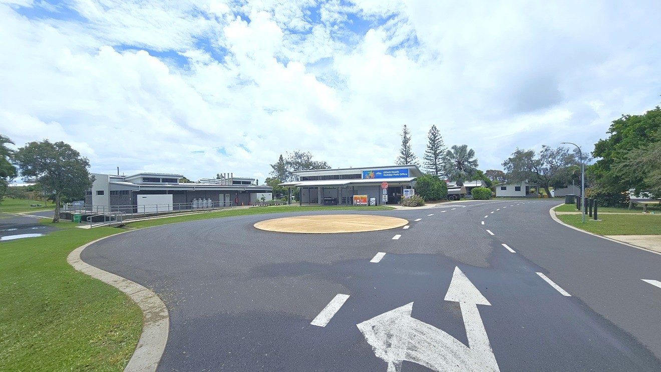 A single-storey building with a flat roof and a sign reading "Elliott Heads Holiday Park Office" is in the centre. The building has an open area with tables and chairs. In front, there are signs including a red "Guests Only Past This Point" and a "10 Shared Zone" sign. The foreground features a grassy area and a paved path leading to the entrance. Trees and additional buildings are visible in the background.