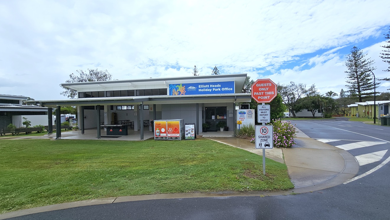 The entrance to the building is a manual glass door opening outward. A black rubber mat and potted plants adorn the front. Posters decorate the walls and door. Inside, the reception desk with a timber front is visible. The interior has wooden floors and a white ceiling with light fixtures. Outside, a tall shrub sits in a concrete planter box.