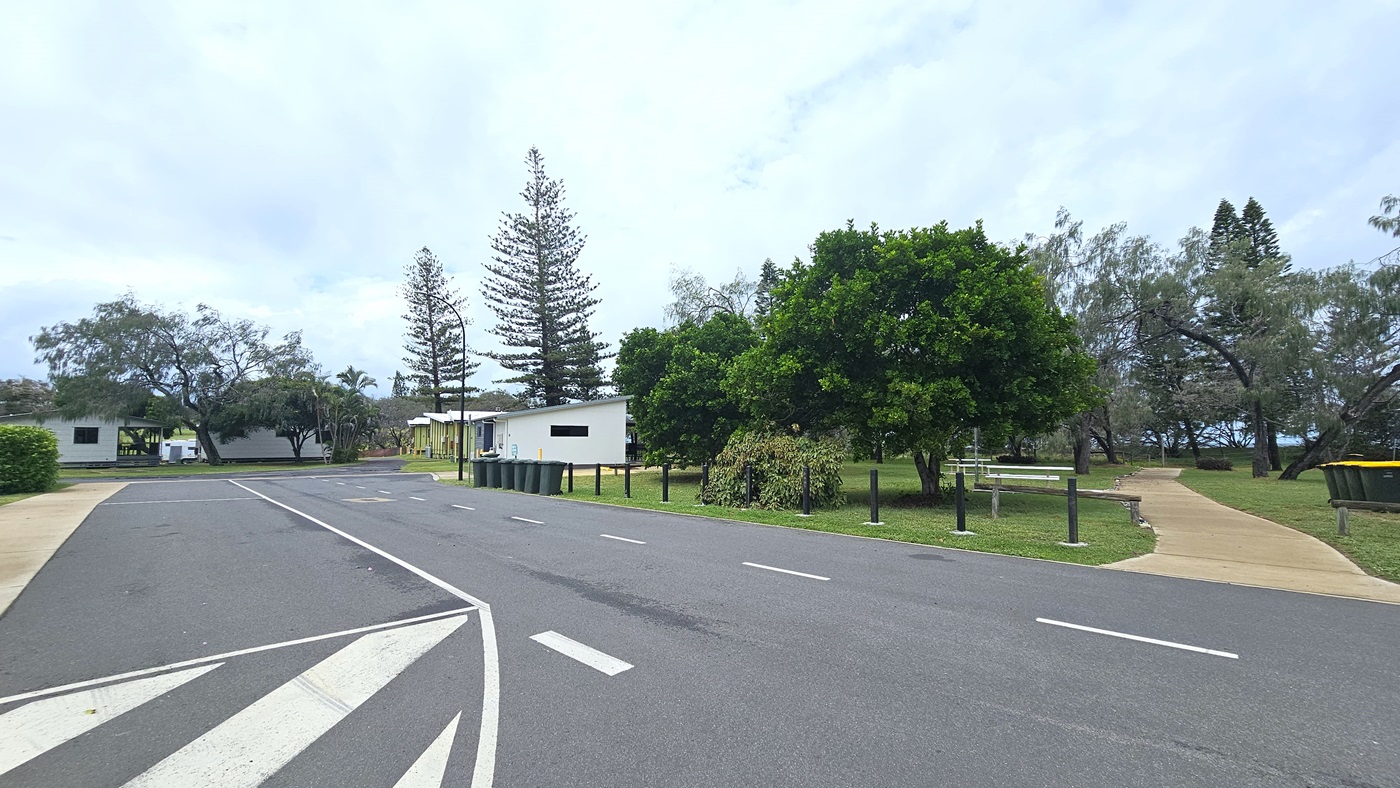 The road features white road markings, bordered by black bollards. A concrete pathway leads to the park on the right, amidst trees and grass. Rubbish bins line the road's edge. In the distance, cabins and a tall street light are visible.