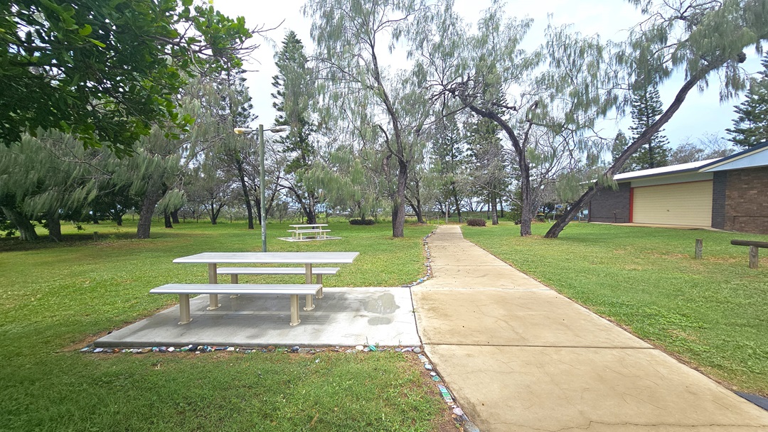 A concrete pathway cuts through grass, leading to picnic tables on the left, set on a concrete floor amidst grass. On the right is a low-rise building with a brown brick facade. Various trees and grass surround the area.