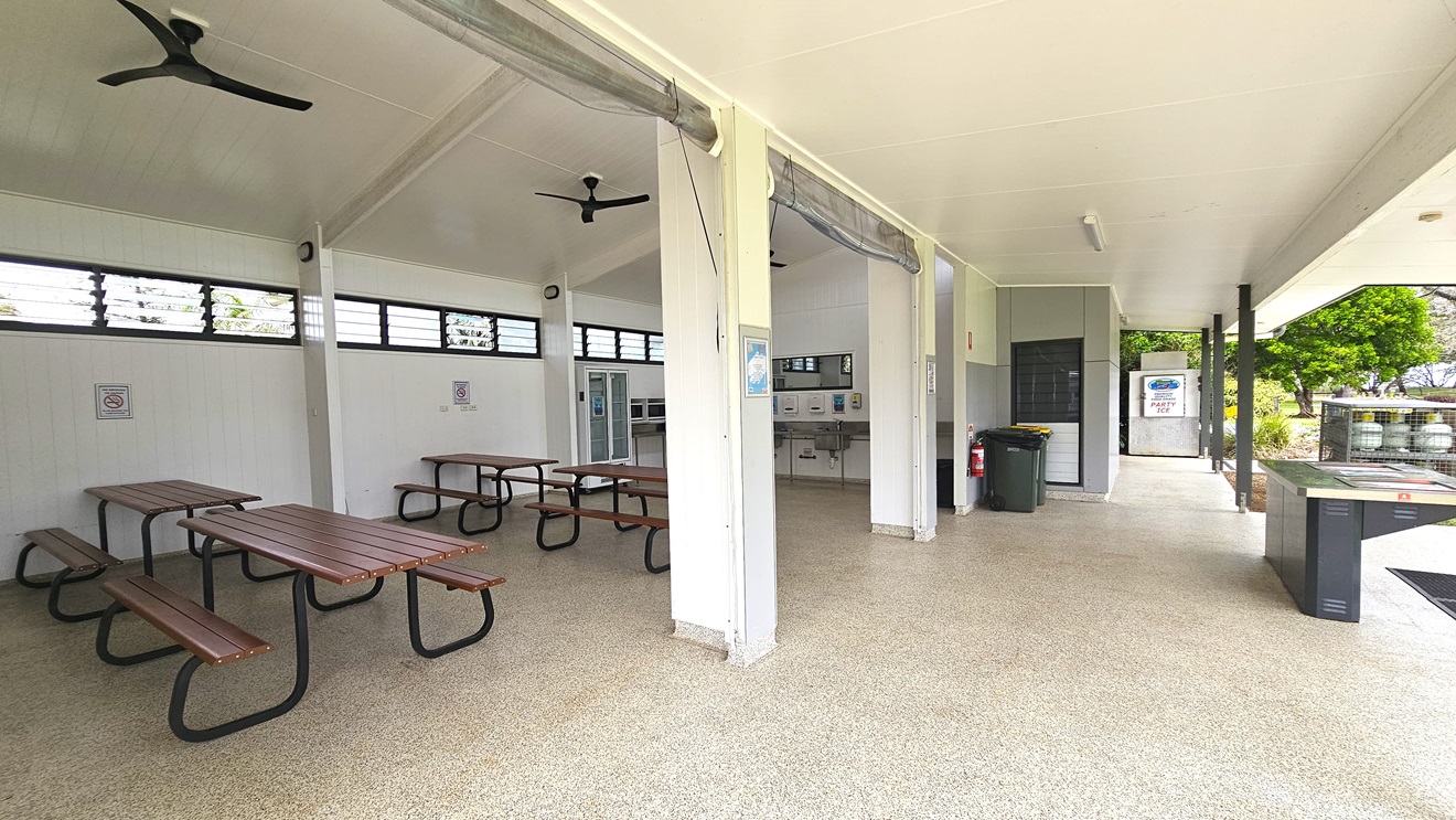 The BBQ area is spacious with picnic tables on the sides and a fridge. A sink and mirror are in the middle wall, and to the right is a barbecue grill. Gas tanks are in a cage nearby, with two rubbish bins and a fire extinguisher on the outer wall. White ceilings and walls feature ceiling fans and jalousy windows for ventilation. Safety reminders are affixed to the walls. Trees are visible on the right.