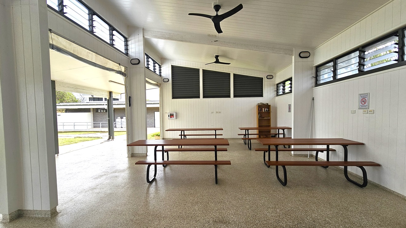 The BBQ area is spacious with picnic tables and a wooden display rack in the right corner. The pitched white ceiling has ceiling fans. The walls feature a white timber design, with jalousy windows and light fixtures on the upper wall. Safety reminders are affixed to walls. To the left is a low-rise facility building nestled on the green grass.