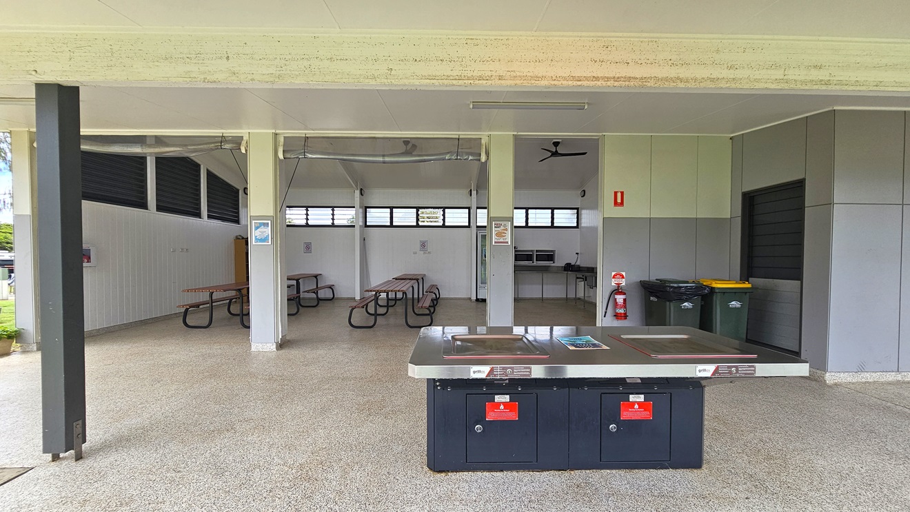 The spacious BBQ area has picnic tables and a wooden display rack in the left corner. On the right, there's a fridge, microwave ovens, and a sink. The pitched white ceiling hosts ceiling fans. Walls showcase a white timber design, with jalousy windows and light fixtures. Safety reminders are affixed to walls. In front is a barbecue grill with safety warning stickers. Two rubbish bins and a fire extinguisher are positioned on the outer wall on the right.
