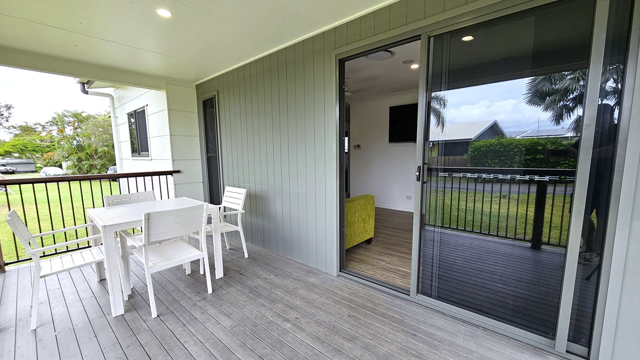 The verandah boasts a grey timber floor with white tables and chairs near metal railings. The wall exhibits a grey wooden plank design, while the ceiling is white. A glass sliding door is located to the right. Inside the cabin, a timber floor is accompanied by a green couch and a flatscreen TV mounted on a white wall. Surrounding the cabin are trees, grass, and visible vehicles.
